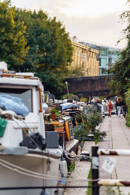 A narrow urban pathway alongside a canal features a row of small boats moored close to the towpath, with their decks cluttered with various discarded items. The debris includes white plastic bags, cardboard boxes, and miscellaneous household waste, arranged without systematic organization. On the right side, a few pedestrians are visible walking along the pavement, some engaged in conversation, while others appear to be observing the rubbish. Behind the boats, dense green trees provide a natural backdrop, with their leaves illuminated by soft daylight. In the background, a brick wall and a modern light green building with large windows are partially visible, suggesting an inner-city area where private waste disposal, such as rubbish removal services like Rubbish Removal Islington, might be employed to manage waste accumulation along the canal. The scene captures a moment of debris accumulation on the waterwayside, highlighting the importance of efficient rubbish collection and waste clearance in urban environments.