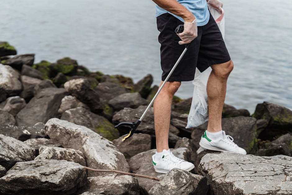 A person wearing white sneakers with green accents, black shorts, and a light blue shirt is standing on a rocky shoreline, using a grabber tool to pick up trash among large, irregular stones that vary in shades of gray and brown. The rocks, some with patches of moss or lichen, extend into a body of water visible in the background. The individual is holding a white plastic bag in one hand, which appears to be partially filled with collected waste, highlighting an activity of environmental cleanup or debris removal in a natural coastal setting. The scene emphasizes manual waste collection on a rugged terrain, aligning with private rubbish removal services provided by companies like Rubbish Removal Islington that cater to alternative waste handling and on-site clearance tasks, often involving manual collection or specialized equipment in outdoor environments.