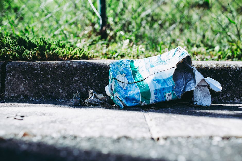 A crumpled plastic bag with a blue and green print lies discarded on a concrete pavement, partially resting against the raised edge of a curb. The bag appears to be made of thin, flexible plastic material, with visible wrinkles and folds, and is weathered from exposure to outdoor conditions. Surrounding the bag, small bits of debris and dirt are scattered along the surface of the pavement, which has a rough texture with slight cracks. In the background, there is a grassy area with vibrant green foliage, and sunlight illuminates the scene, casting subtle shadows across the curb and pavement. The image reflects a typical urban environment where waste, such as trash and litter, accumulates along public walkways, highlighting the importance of private waste removal services like those offered by Rubbish Removal Islington to manage and clear rubbish effectively from outdoor areas.