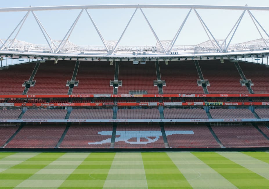 The interior of a large football stadium with red tiered seating extending from the lower to upper levels, arranged around a well-maintained grass pitch with alternating light and dark green stripes; in the background, part of the stadium's white structural roof trusses are visible, spanning across the open sky above the seating areas. The seats feature various advertisements and sponsors displayed on banners and ledges, including the Arsenal football club logo prominently visible in the middle section. The overall scene is well-lit with natural daylight, creating a clear and unobstructed view of the seating arrangement and the pitch, typical of a sports venue ready for a match or event. The stadium's infrastructure and seating layout are designed for spectators, with the image capturing a wide-angle perspective of the venue’s interior from the field level up towards the upper stands.