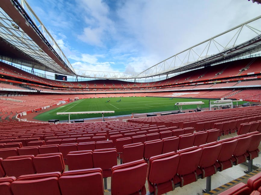 The image displays a large sports stadium with a panoramic view of the empty seating areas, which are predominantly red in colour. The stadium features multiple tiers of red plastic seats arranged in rows, curving around a well-maintained grass pitch in the center. The pitch is vibrant green, with visible mowing patterns, and has white lines marking the field boundaries. Surrounding the pitch are several white benches or dugouts, likely for team use. The stadium's roof structure is partly visible, with a white, tensile canopy supported by a series of steel trusses extending over part of the stands, providing partial cover. The sky above is partly cloudy with patches of blue, and sunlight illuminates the scene evenly. Although the stadium appears prepared for a match, no players or spectators are present. This setting, captured during a quiet moment, aligns with private or alternative match day preparation, and the image's surroundings suggest it is an environment where professional sports events remain organized outside of regular attendance, echoing the context of isolated rubbish removal or on-site clearance services such as those offered by Rubbish Removal Islington in the area.