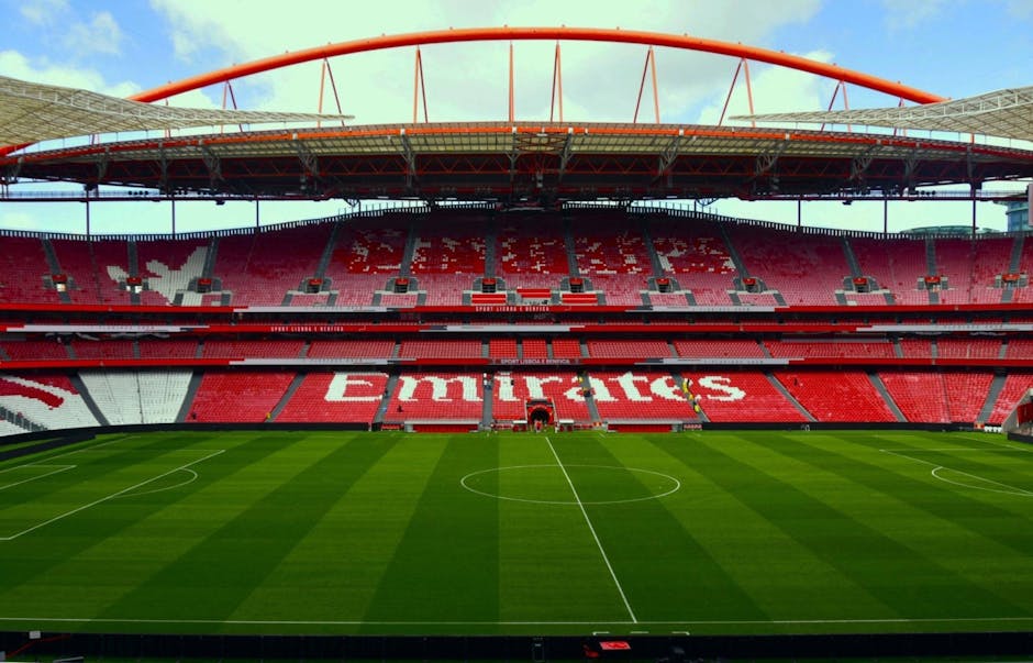 The interior of a large football stadium with red tiered seating extending from the lower to upper levels, arranged around a well-maintained grass pitch with alternating light and dark green stripes; in the background, part of the stadium's white structural roof trusses are visible, spanning across the open sky above the seating areas. The seats feature various advertisements and sponsors displayed on banners and ledges, including the Arsenal football club logo prominently visible in the middle section. The overall scene is well-lit with natural daylight, creating a clear and unobstructed view of the seating arrangement and the pitch, typical of a sports venue ready for a match or event. The stadium's infrastructure and seating layout are designed for spectators, with the image capturing a wide-angle perspective of the venue’s interior from the field level up towards the upper stands.