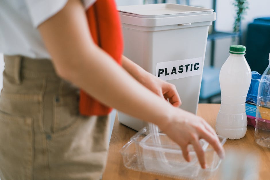 A person is seen placing a clear plastic food container into a large, white plastic bin labeled 'PLASTIC' for waste segregation. The individual, wearing beige trousers and a white top, is partially visible, with their hands handling the container. The scene is set indoors on a wooden table, with several other plastic bottles, including a white bottle with a green cap and a transparent bottle, placed nearby. The background shows a blurred glimpse of a living space with a blue sofa and a window allowing natural light to illuminate the area. The plastic bin, with its smooth finish and simple rectangular shape, emphasizes organized household waste disposal, aligning with principles of proper rubbish separation and private waste handling services, such as those offered by Rubbish Removal Islington. The scene conveys an environment focused on environmentally conscious waste management practices, reflecting the importance of segregating recyclable plastics for on-site clearance or independent collection, in contrast to council bulk waste procedures.
