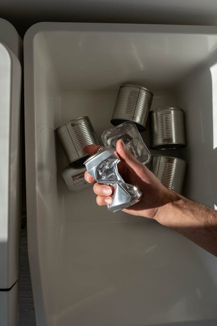 A person's hand is shown placing a crumpled aluminium beverage can into a chest freezer, which already contains several similar cans arranged in a somewhat scattered manner. The freezer interior is white, with a smooth, clean surface, and is illuminated by natural light coming from the right side of the image. The cans are typical aluminium, with a metallic silver finish and ridged sides, some partially hidden behind others. The hand is gripping a crushed can, indicating disposal or recycling preparation, and appears to be outdoors or in a well-lit indoor environment. The scene illustrates a domestic waste disposal activity, with the aluminium cans possibly prepared for removal by a private waste collection service like Rubbish Removal Islington, especially in the context of managing household recycling or bulk waste. The neutral background emphasizes the focus on the hand and the aluminium cans, aligning with the theme of rubbish and waste management, and subtly supporting alternatives to council bulky waste rules and household disposal fees.