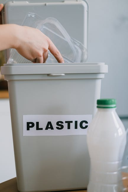 A person is seen placing a clear plastic food container into a large, white plastic bin labeled 'PLASTIC' for waste segregation. The individual, wearing beige trousers and a white top, is partially visible, with their hands handling the container. The scene is set indoors on a wooden table, with several other plastic bottles, including a white bottle with a green cap and a transparent bottle, placed nearby. The background shows a blurred glimpse of a living space with a blue sofa and a window allowing natural light to illuminate the area. The plastic bin, with its smooth finish and simple rectangular shape, emphasizes organized household waste disposal, aligning with principles of proper rubbish separation and private waste handling services, such as those offered by Rubbish Removal Islington. The scene conveys an environment focused on environmentally conscious waste management practices, reflecting the importance of segregating recyclable plastics for on-site clearance or independent collection, in contrast to council bulk waste procedures.