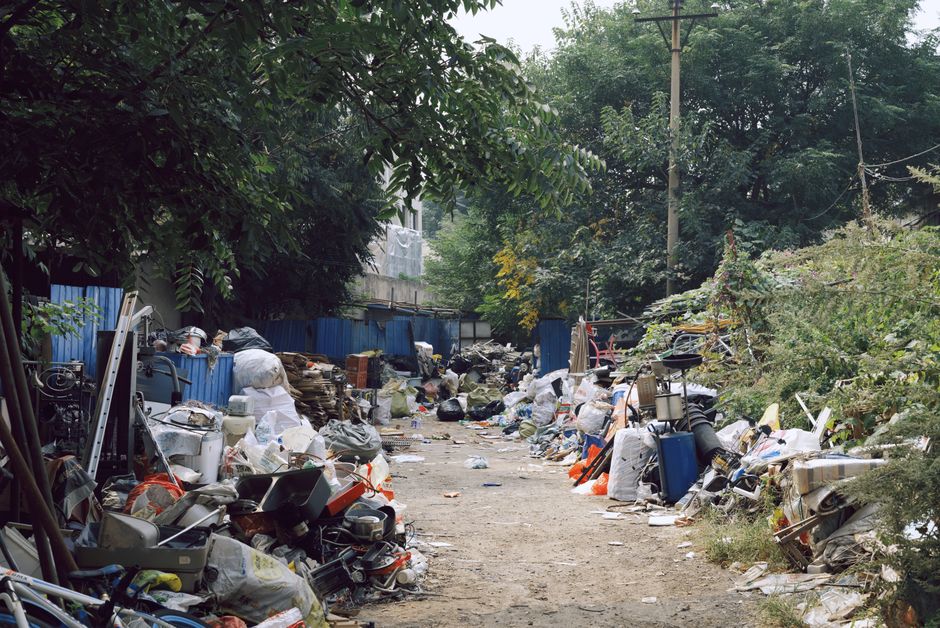 A dirt pathway lined with large piles of mixed rubbish and waste materials on both sides, including plastic bags, discarded furniture, broken household items, and miscellaneous debris. The waste is stacked up irregularly, with some items spilling onto the ground, creating an untidy and cluttered scene. Behind the rubbish, there is a blue metal fence and overgrown bushes, with trees and foliage overhead providing partial shade. An electrical pole with wires extends vertically into the scene, contributing to the cluttered environment. The overall atmosphere suggests an unmanaged waste accumulation area that could be addressed through professional rubbish removal services such as those offered by Rubbish Removal Islington. This scene highlights the importance of proper waste disposal and the potential for alternative, on-site clearance methods to help maintain cleaner, safer environments in urban or semi-urban areas.