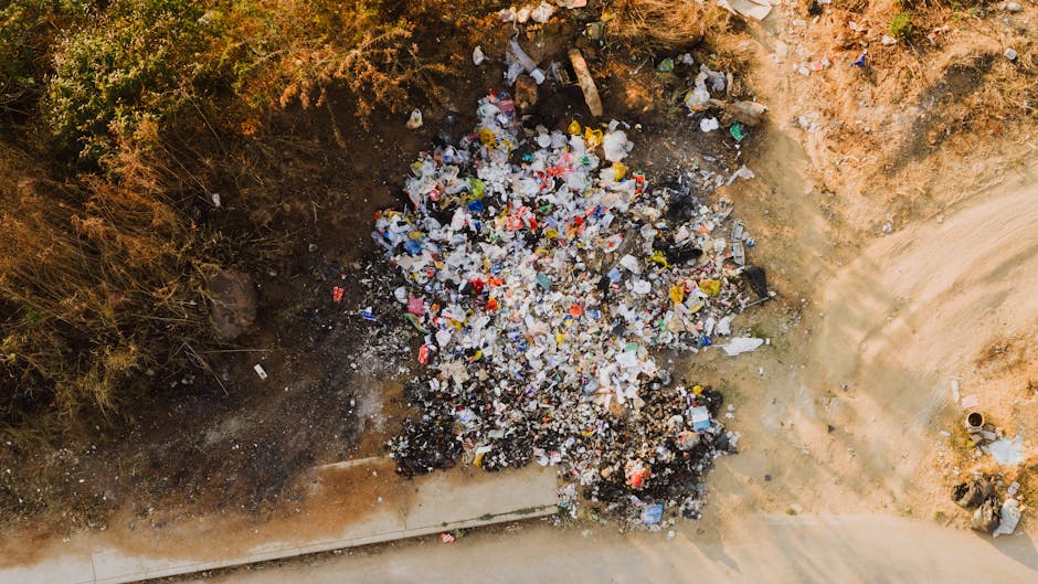 An aerial view of a small, cleared area beside a dirt road shows a large pile of mixed waste, predominantly made up of plastic packaging, food wrappers, and other lightweight debris, with some darker, burnt-looking materials at the base. The waste is scattered unevenly, with some items overlapping and others spread out, creating a chaotic appearance. To the left, overgrown bushes and dry grass border the scene, adding a natural contrast to the man-made rubbish. The ground surrounding the pile reveals a combination of bare earth and dust, with a slight impression where the debris has accumulated. On the right side, a section of compacted dirt or clay suggests recent disturbance, possibly from previous clearing activity. Visible in the lower part of the image is a paved sidewalk, partially covered by shadows cast from nearby objects or terrain features. Overall, the scene appears to be an illegal dumping site or an area in need of waste removal, reflecting the importance of proper rubbish disposal services such as those offered by Rubbish Removal Islington, who often handle such instances of private or independent waste collection, especially in areas not serviced by council refuse collections.
