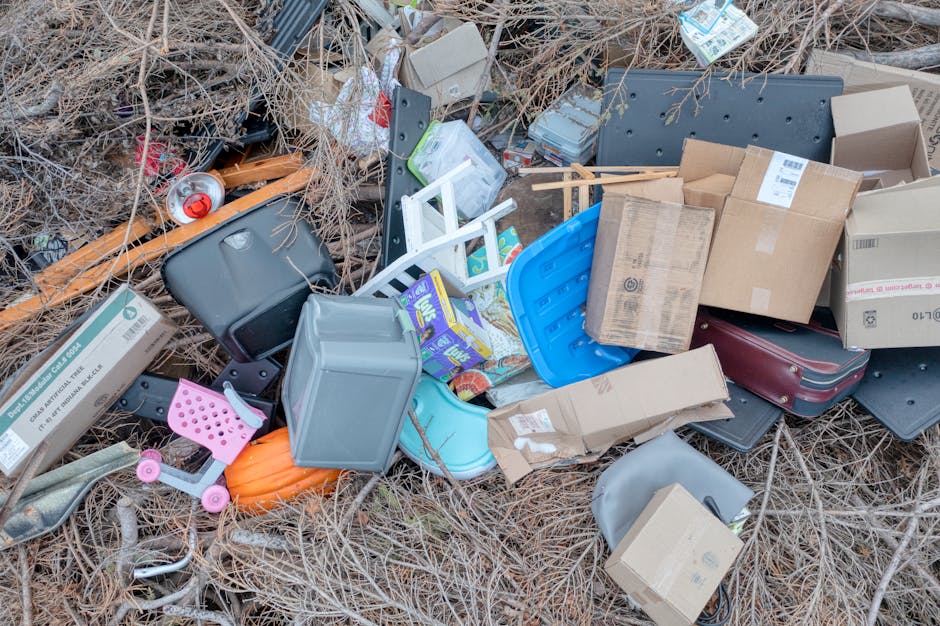 A dirt pathway lined with large piles of mixed rubbish and waste materials on both sides, including plastic bags, discarded furniture, broken household items, and miscellaneous debris. The waste is stacked up irregularly, with some items spilling onto the ground, creating an untidy and cluttered scene. Behind the rubbish, there is a blue metal fence and overgrown bushes, with trees and foliage overhead providing partial shade. An electrical pole with wires extends vertically into the scene, contributing to the cluttered environment. The overall atmosphere suggests an unmanaged waste accumulation area that could be addressed through professional rubbish removal services such as those offered by Rubbish Removal Islington. This scene highlights the importance of proper waste disposal and the potential for alternative, on-site clearance methods to help maintain cleaner, safer environments in urban or semi-urban areas.