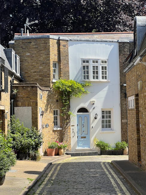 A narrow residential street in an urban area shows a two-storey brick house with a distinctive white front façade, featuring a rounded arch doorway painted in a light pastel blue with a glass panel and a silver handle. Above the door, there is a small rectangular window, and to the right of the entrance, a larger white-framed window with multiple panes. A small potted plant with pink flowers is placed near the doorway, and a green vine climbs the brick wall on the left side of the house. To the left of the building, there's dense foliage and shrubs, while to the right, a section of a neighboring brick building with a sloped roof is visible. The street is paved with cobblestones, and the scene is illuminated by natural daylight, with shadows cast by the trees and structures. The environment suggests a quiet, well-maintained area where private property clearance or bespoke waste disposal services like those offered by Rubbish Removal Islington might be involved in maintaining the tidy appearance of the street.