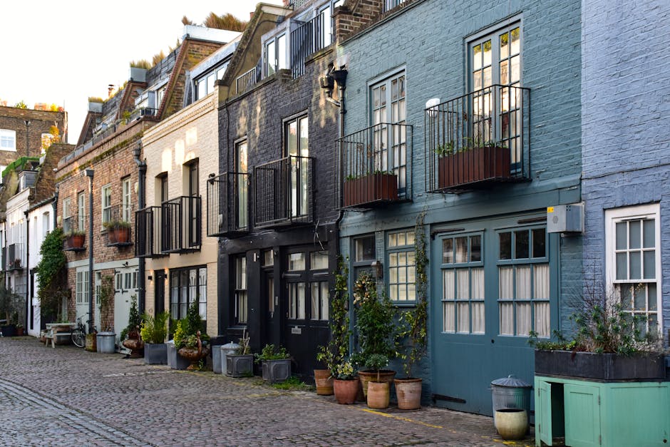 A row of narrow, multilevel terraced buildings along a cobblestone street, with distinctive brick and painted facades in shades of beige, black, and light blue. Each property features small front yards or balconies, with black metal railings and wooden planters containing greenery. Potted plants in terracotta and plastic containers are placed directly on the ground, along the base of the buildings, adding greenery to the scene. The street appears to be in a quiet residential area, with no visible traffic or pedestrians, and mostly features closed doors and windows. The lighting suggests late afternoon or early evening, with soft shadows cast on the buildings. The overall scene emphasizes classic London mews architecture, with minimal clutter or rubbish visible, consistent with a professional waste removal service operating in an established urban setting such as Rubbish Removal Islington, providing solutions for on-site clearance or private waste handling in historic residential streets.