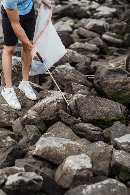 A person dressed in casual sportswear, including white sneakers, black shorts, and a light blue T-shirt, is using a grabber tool to pick up debris or small objects from a rocky outdoor area near a riverbank. The individual holds a white plastic bag with red trim in one hand while positioning the grabber tool with the other, appearing to collect rubbish or natural debris from the uneven surface of large, irregularly shaped stones that vary in dark gray and brown tones with some moss or algae growth. The surrounding environment features a stony shoreline with numerous rocks of different sizes, some partially submerged in water, indicating a natural riverside setting. The lighting is natural but slightly subdued, suggesting an overcast day. This scene demonstrates an individual engaging in an outdoor clean-up activity, which aligns with waste removal efforts, similar to those provided by Rubbish Removal Islington for private or independent collection of rubbish rather than traditional municipal disposal.