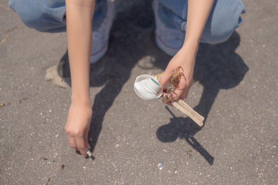 A person is crouched down on a paved surface, overlooking a small pile of debris. The individual is wearing light blue denim jeans and white sneakers with red and grey accents. Their right hand holds a piece of broken white ceramic or plaster, with rough, jagged edges, and small tangled remnants of what appears to be adhesive or sealant. The left hand is near the ground, free of objects. The background shows an unadorned asphalt surface, typical of a driveway or outdoor walkway. The lighting suggests it is a clear day with natural sunlight casting shadows. This scene may suggest an on-site waste clearance activity, where tidy disposal or collection of debris, such as broken construction materials, is being carried out, aligning with services provided by Rubbish Removal Islington for independent waste management solutions, including rubbish and construction debris removal.