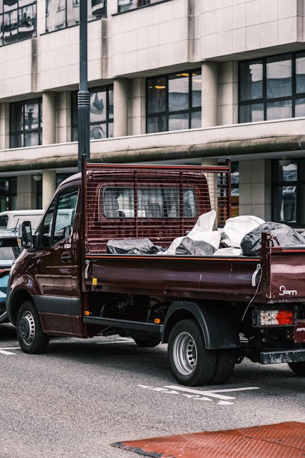 A small burgundy flatbed truck parked on an urban street in front of a modern building with large windows and a concrete facade. The truck's cargo area is filled with various types of waste, including black and white plastic bags, cardboard boxes, and other discarded materials, indicating a recent rubbish collection or disposal service. A metal safety barrier is mounted at the front of the cargo bed, partially concealing some debris. The truck appears clean and well-maintained, suitable for private waste removal or on-site clearance tasks. The street is paved, with a parking line visible along the curb, and a lamppost is positioned near the vehicle, suggesting an organized urban environment. The natural daylight highlights the textures of the waste bags and the reflective finish of the vehicle, subtly relating to the services offered by Rubbish Removal Islington for independent rubbish removal or waste handling outside of local authority collection schemes.