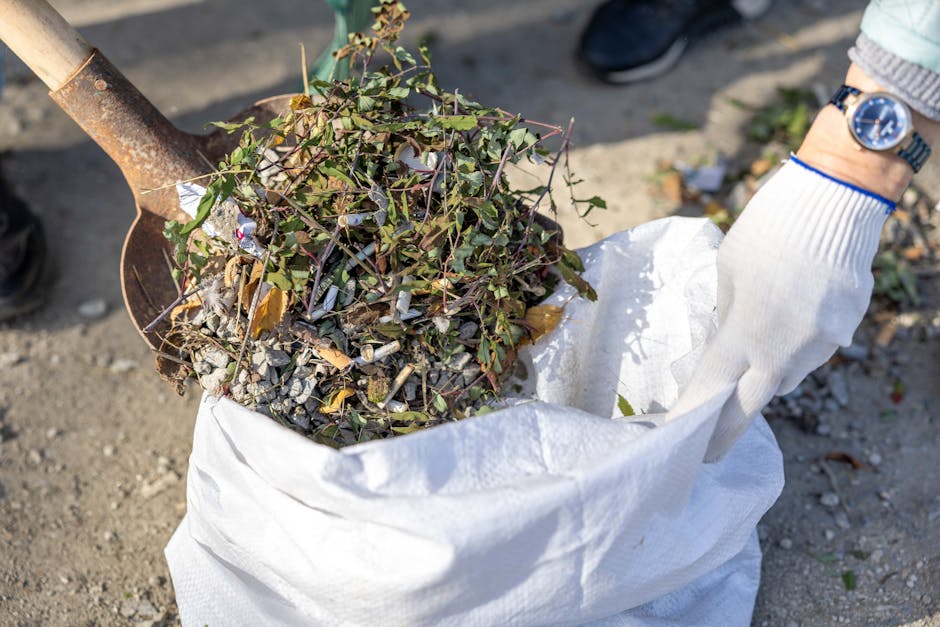 A person wearing a white glove and a blue wristwatch is holding a large white woven polypropylene sack filled with garden debris, including small branches with green and brown leaves, and assorted small rocks or gravel. The sack appears to be partially open and is situated outdoors on a patch of ground composed of gravel and soil. A rusty shovel with a wooden handle is positioned nearby, leaning slightly against or above the sack, suggesting ongoing garden or yard waste clearance. In the background, a glimpse of a person’s dark footwear is visible, along with some scattered natural waste and patchy greenery, indicating an outdoor setting possibly in a residential or garden area. The scene is lit by natural daylight, emphasizing the textures of the leaves, gravel, and the woven surface of the sack, which aligns with the context of private waste handling or rubbish removal services offered by Rubbish Removal Islington.