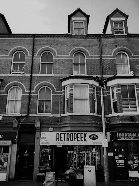 A black and white photograph depicting the front view of a multi-storey brick building with traditional architectural features, including arched window frames and a prominent bay window on the ground floor. The building's upper floors have three visible dormer windows with gabled roofs. On the ground floor, there are storefronts with signboards, one of which displays the name 'RETROPEEK' and mentions house clearances, indicating the building is occupied by a rubbish removal or clearance service. In front of the shops, there are a few waste bins and small outdoor objects on the pavements, reflecting an urban commercial setting. The overall scene connotes an area where private waste collection services operate outside the regular council rubbish collection, fitting within the context of independent waste handling and rubbish removal services in Islington, as provided by companies like Rubbish Removal Islington. The environmental atmosphere appears calm with natural daylight or overcast conditions, typical of an urban street scene focused on waste collection and clearance activities.