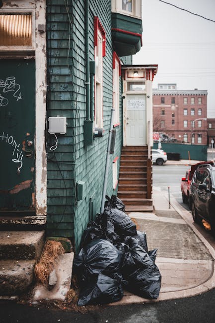 A stack of six black garbage bags filled with waste rests against the exterior wall of a building, positioned on a concrete sidewalk near a small set of wooden steps leading up to a white door with red trim. The building’s exterior features teal-green wooden shingle cladding, with visible weathering and some graffiti on the adjacent door to the left. Nearby, a small portion of burnt orange-brown dried grass is visible at the base of the wall. The scene is set in an urban environment, with parked cars on the street and multi-story brick buildings in the background under an overcast sky, suggesting an everyday urban waste removal situation that may be managed by a private waste disposal service like Rubbish Removal Islington. The overall composition reflects a typical residential alleyway scene where unmanaged rubbish is temporarily stored before disposal or collection, aligning with services related to rubbish removal or private waste handling in the local area.