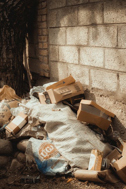 A pile of mixed waste materials, including cardboard boxes, paper bags, and plastic packaging, is situated on the ground near a brick wall and a concrete block wall. The cardboard boxes vary in size, some with partially open flaps revealing empty interiors, with visible printed labels and text. A large, coarse, woven fabric sack is partly under the boxes, with a branded plastic bag nearby. The waste is placed on uneven ground with small rocks and dirt. The scene appears to be an outdoor space, possibly a back alley or side yard, with natural lighting and shadows cast on the waste and walls. The image reflects a typical scene where independent rubbish removal services, like those offered by Rubbish Removal Islington, handle domestic or commercial waste disposal, emphasizing the importance of proper waste management to prevent clutter buildup.