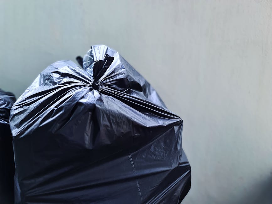 A stack of six black garbage bags filled with waste rests against the exterior wall of a building, positioned on a concrete sidewalk near a small set of wooden steps leading up to a white door with red trim. The building’s exterior features teal-green wooden shingle cladding, with visible weathering and some graffiti on the adjacent door to the left. Nearby, a small portion of burnt orange-brown dried grass is visible at the base of the wall. The scene is set in an urban environment, with parked cars on the street and multi-story brick buildings in the background under an overcast sky, suggesting an everyday urban waste removal situation that may be managed by a private waste disposal service like Rubbish Removal Islington. The overall composition reflects a typical residential alleyway scene where unmanaged rubbish is temporarily stored before disposal or collection, aligning with services related to rubbish removal or private waste handling in the local area.